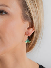 A close-up of a woman's ear wearing a small dangling earring with a green and blue fan-shaped charm on a gold hook, against a light gray background.