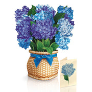 Bouquet of blue flowers in a woven basket with a blue ribbon, accompanied by a matching card on a white background.