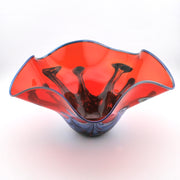 A hand-blown glass bowl with a wavy, flared rim, featuring a vibrant red-orange outer layer and dark purple-black internal swirls, set against a plain white background.