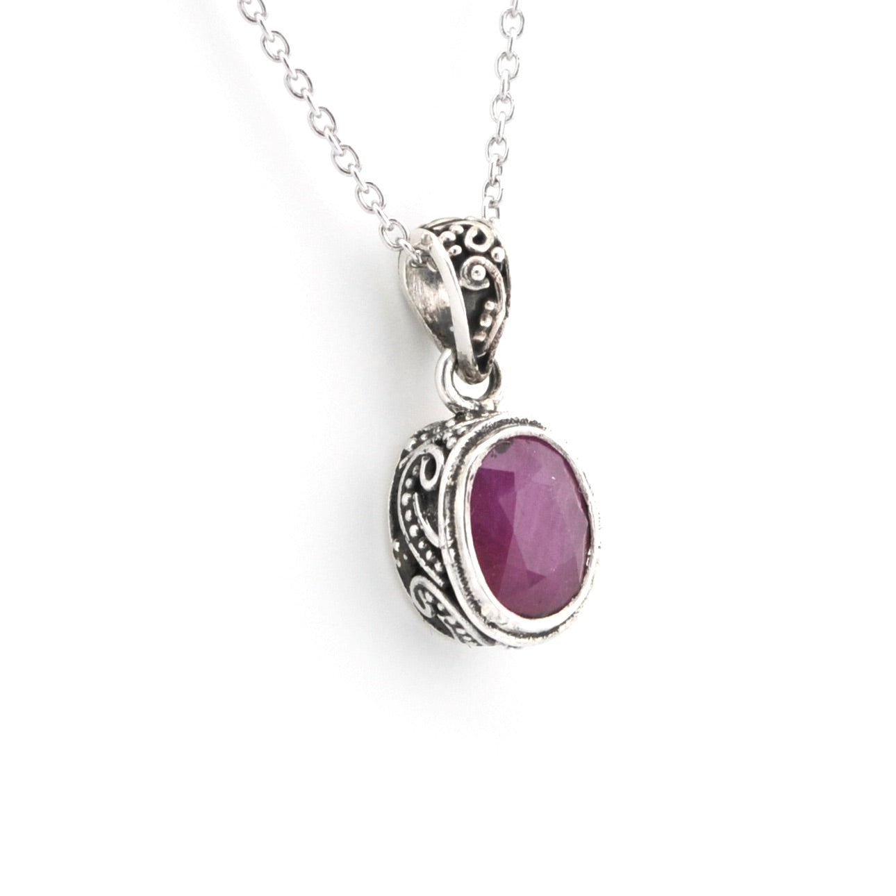 A silver pendant necklace featuring an oval purple gemstone in an ornate filigree bezel setting, suspended from a delicate silver chain against a plain white background.