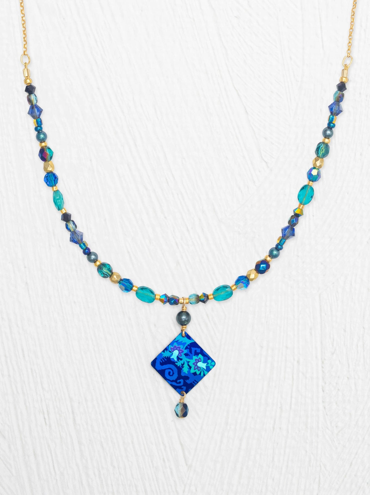 A beaded necklace with a central blue patterned diamond pendant, featuring turquoise, teal, and blue beads with gold-toned metal links and spacer beads, displayed against a white textured background.
