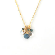 A delicate gold chain necklace featuring a cluster pendant with a large blue stone, smaller green and gray stones, and gold wire accents, set against a plain white background.
