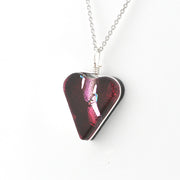 A heart-shaped glass pendant necklace with an iridescent dark red and magenta finish, suspended from a silver chain on a white background.