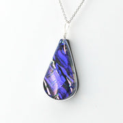 A teardrop-shaped pendant necklace featuring a vibrant, iridescent blue and purple dichroic glass set in a silver-toned metal frame, suspended from a silver chain against a plain white background.