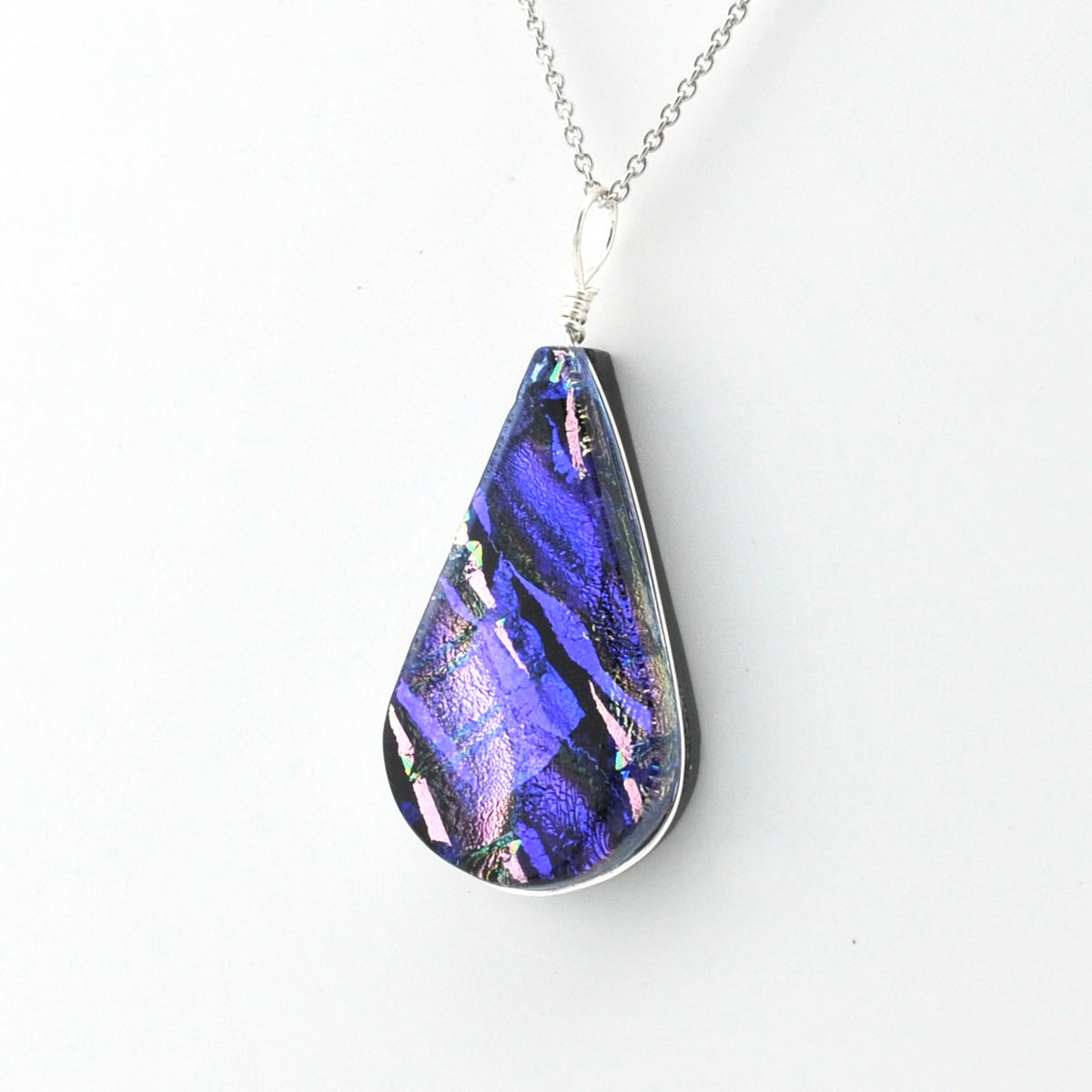 A teardrop-shaped pendant necklace featuring a vibrant, iridescent blue and purple dichroic glass set in a silver-toned metal frame, suspended from a silver chain against a plain white background.