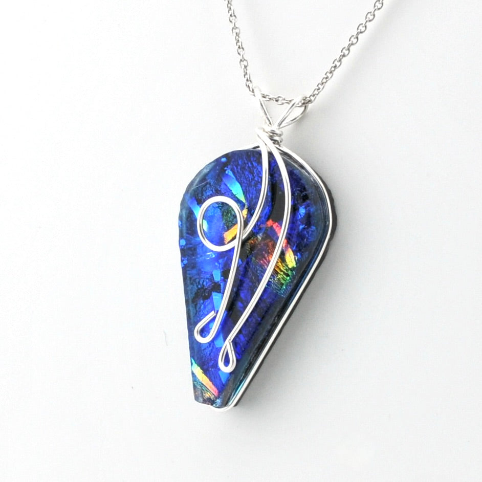 A silver chain necklace with a teardrop-shaped pendant featuring a vibrant blue iridescent dichroic glass and a white cursive monogram design, shown on a plain white background.