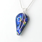 A silver chain necklace with a teardrop-shaped pendant featuring a vibrant blue iridescent dichroic glass and a white cursive monogram design, shown on a plain white background.