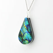 A teardrop-shaped pendant necklace featuring a colorful abstract pattern of green, blue, and yellow on a black background, suspended from a silver chain against a plain white background.