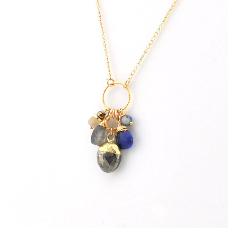A gold chain necklace with a circular ring pendant holding several small gemstone charms, including a prominent dark gray labradorite and a deep blue bead, shown on a plain white background.