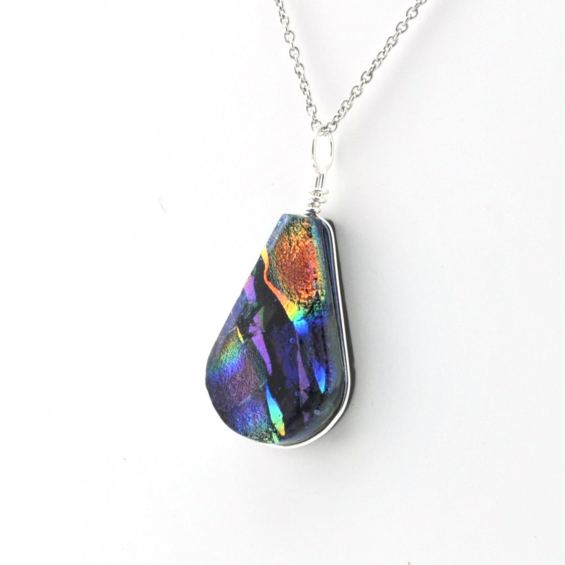 A teardrop-shaped pendant necklace featuring a highly iridescent, dichroic glass piece that shifts between blue, green, orange, and purple, set in a silver bezel and suspended from a silver chain against a plain white background.