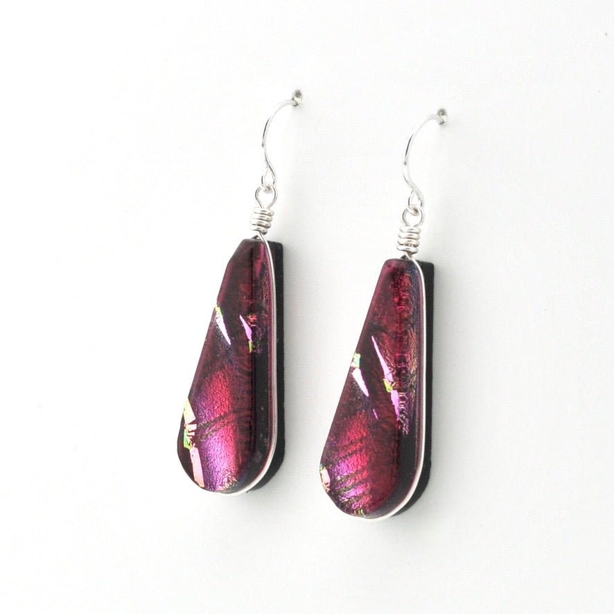 A pair of teardrop-shaped dangle earrings made from dichroic glass in shades of magenta, purple, and pink, suspended from silver fishhook wires on a plain white background.