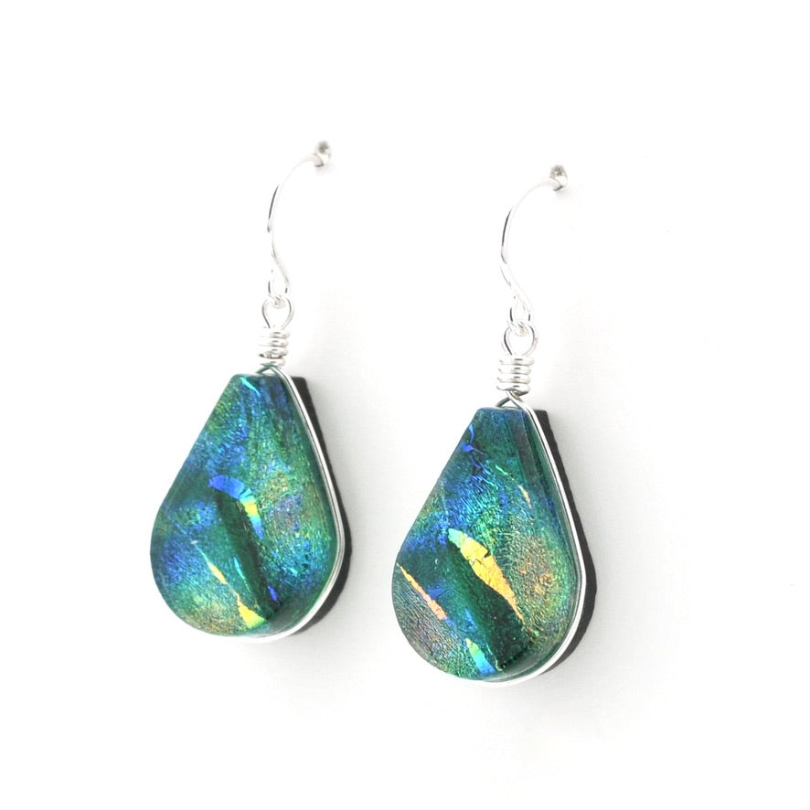 A pair of teardrop-shaped dangle earrings with iridescent blue, green, and gold dichroic glass suspended from silver fishhook wires on a white background.