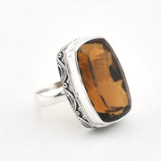 A silver ring featuring a large, cushion-cut brown smoky quartz gemstone, set in an ornate, engraved sterling silver band against a plain white background.