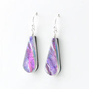 A pair of teardrop-shaped dangle earrings with iridescent purple, pink, and blue dichroic glass, set in silver and suspended from silver fishhook wires, shown against a plain white background.