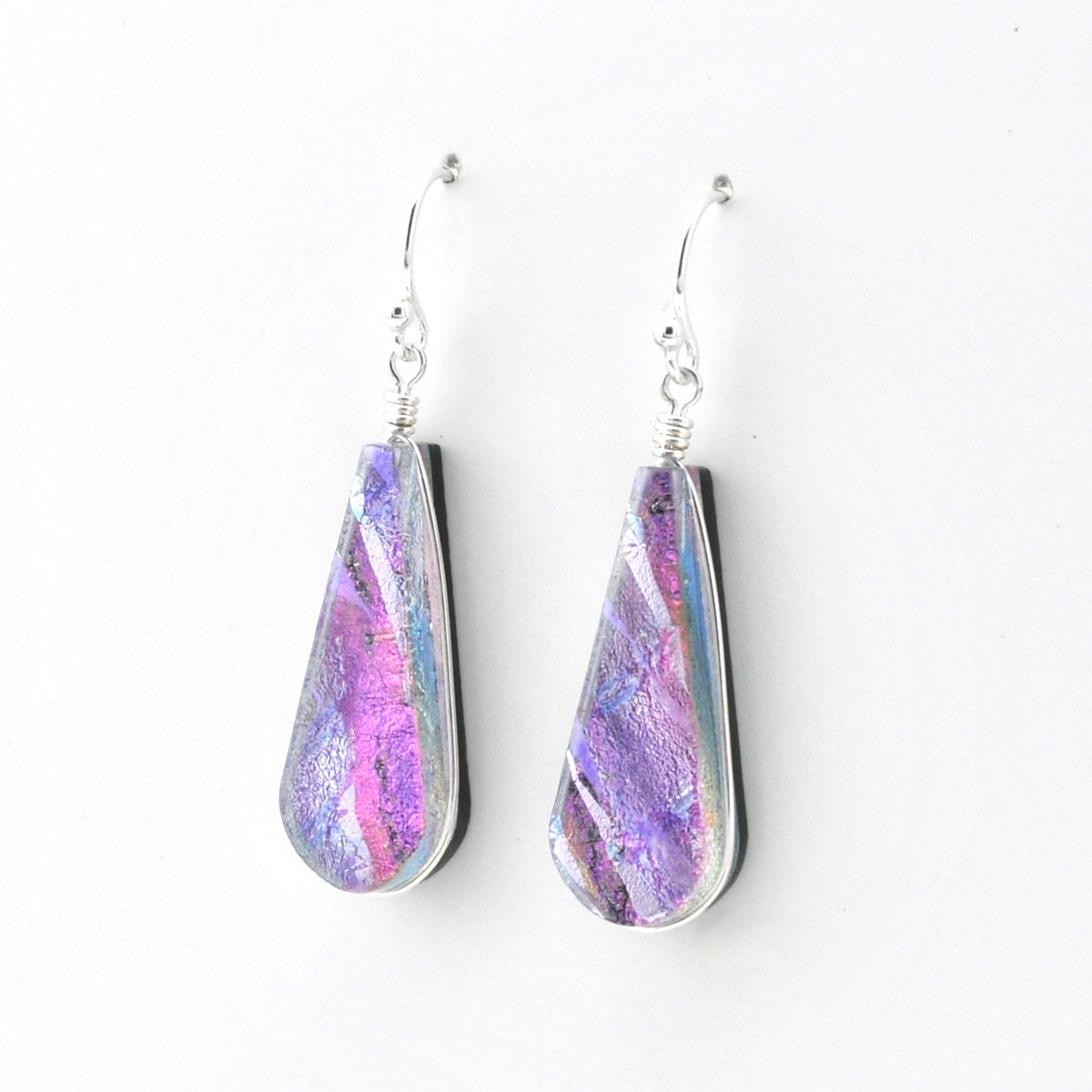 A pair of teardrop-shaped dangle earrings with iridescent purple, pink, and blue dichroic glass, set in silver and suspended from silver fishhook wires, shown against a plain white background.