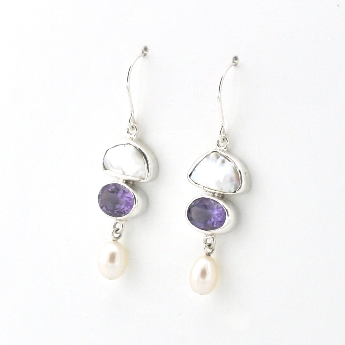 A pair of silver drop earrings featuring a white pearl, a purple amethyst, and a white freshwater pearl dangling below, set against a plain white background.