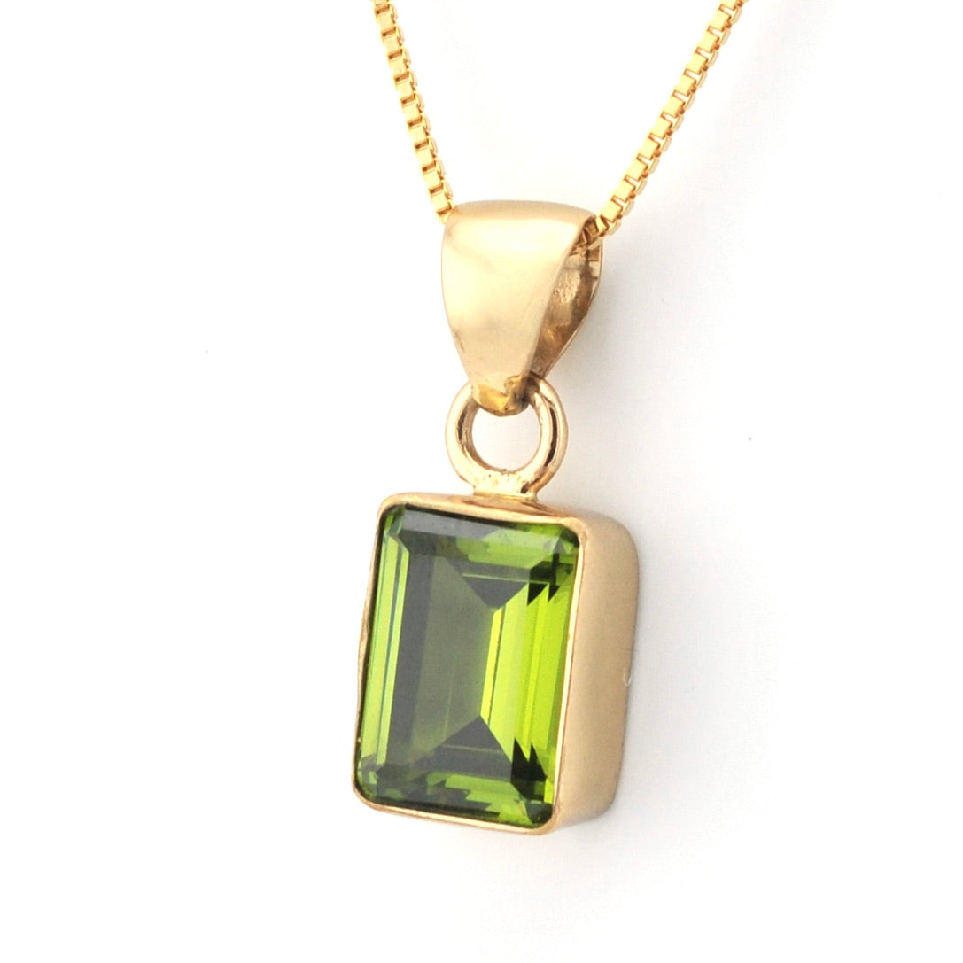 A gold pendant necklace featuring a rectangular green gemstone with a faceted emerald cut, suspended from a gold chain on a white background.