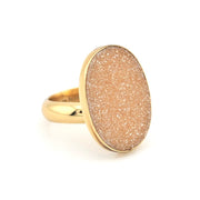 A gold ring featuring a large, oval-shaped dome stone with a textured, glittery champagne-gold finish, set against a plain white background.
