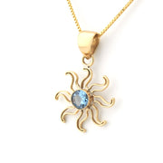 A gold pendant necklace featuring a stylized sun design with a central light blue gemstone, suspended from a gold chain on a white background.