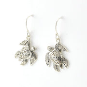 A pair of silver-toned fishhook earrings with detailed sea turtle charms featuring ornate filigree patterns, shown on a plain white background.