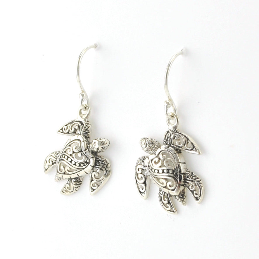 A pair of silver-toned fishhook earrings with detailed sea turtle charms featuring ornate filigree patterns, shown on a plain white background.