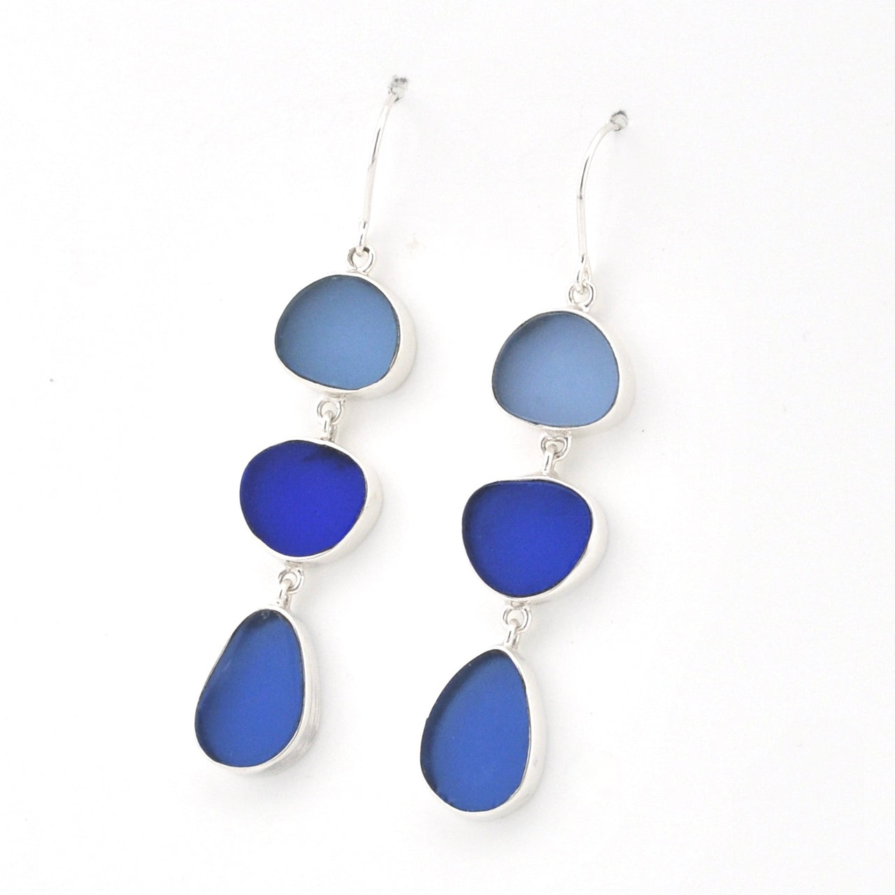 A pair of silver drop earrings featuring three stacked, organic-shaped sea glass pieces in shades of blue, photographed on a clean white background.