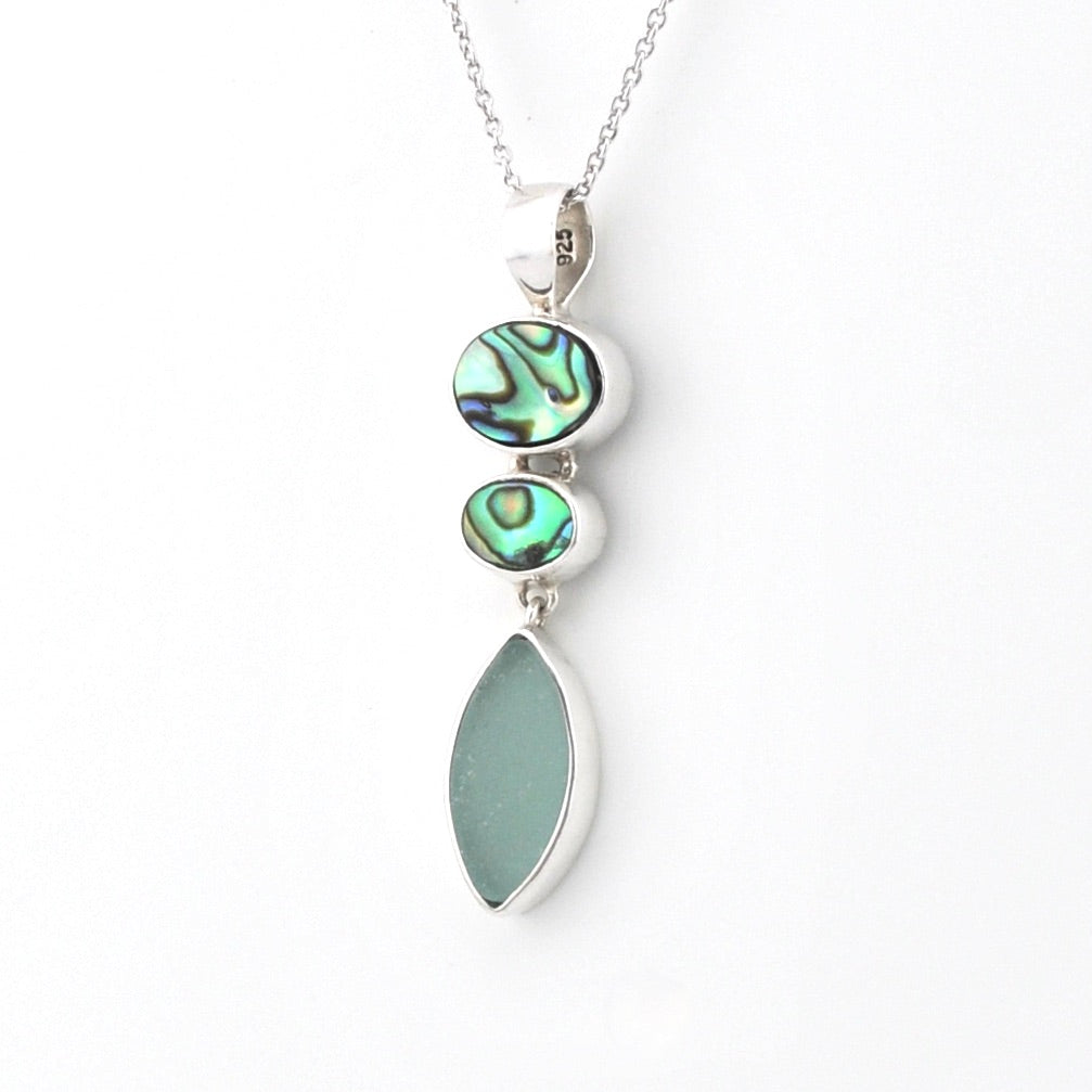 A silver pendant necklace featuring three stacked vertical elements: a large teardrop-shaped light green stone, a smaller oval iridescent abalone shell, and a medium oval abalone shell, suspended from a delicate silver chain on a white background.
