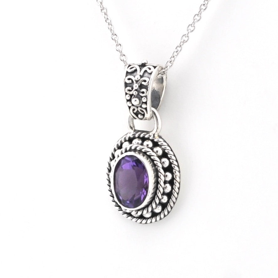 A silver-toned oval pendant necklace featuring a central purple amethyst gemstone, detailed with textured scrollwork and rope-like borders, shown on a plain white background.