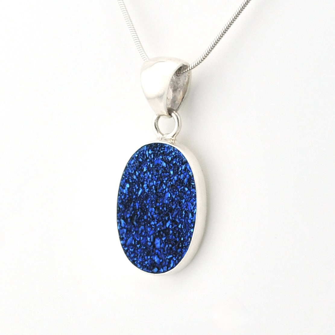 A silver pendant necklace featuring an oval blue glitter druzy stone in a silver bezel setting, suspended from a silver chain against a plain white background.