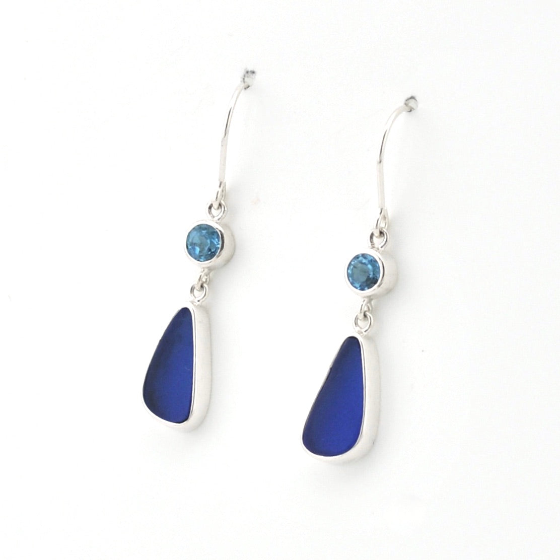 A pair of silver dangle earrings featuring a small round light blue gemstone and a larger teardrop-shaped deep blue sea glass shown on a plain white background.