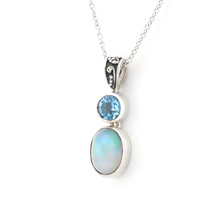A silver pendant necklace featuring a round light blue gemstone and an oval iridescent white opal stone, suspended from a delicate silver chain on a white background.