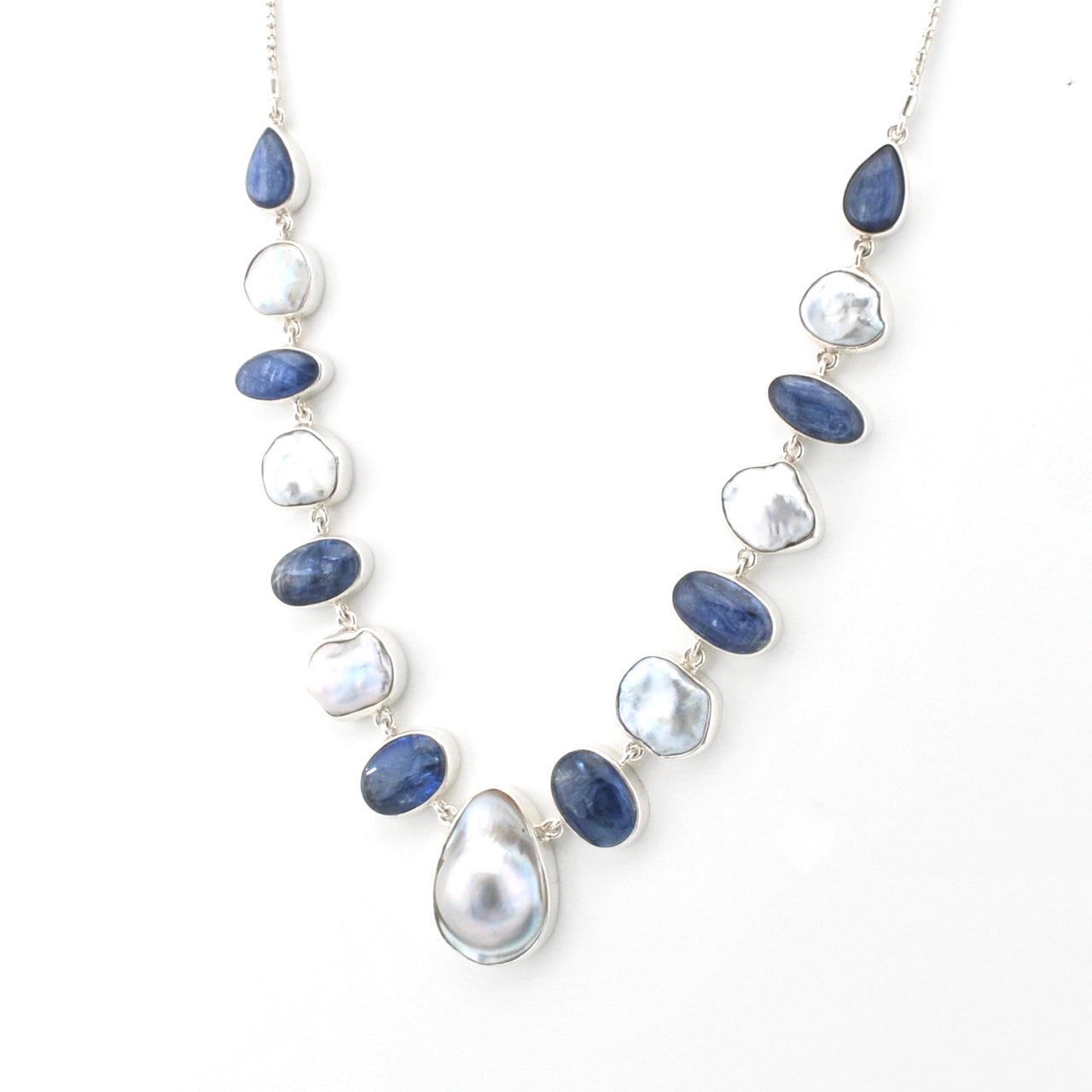 A silver-toned necklace featuring alternating polished dark blue sapphire-like beads and irregular white keshi pearl beads, with a large teardrop-shaped white pearl pendant at the center, displayed on a clean white background.