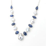 A silver-toned necklace featuring alternating polished dark blue sapphire-like beads and irregular white keshi pearl beads, with a large teardrop-shaped white pearl pendant at the center, displayed on a clean white background.