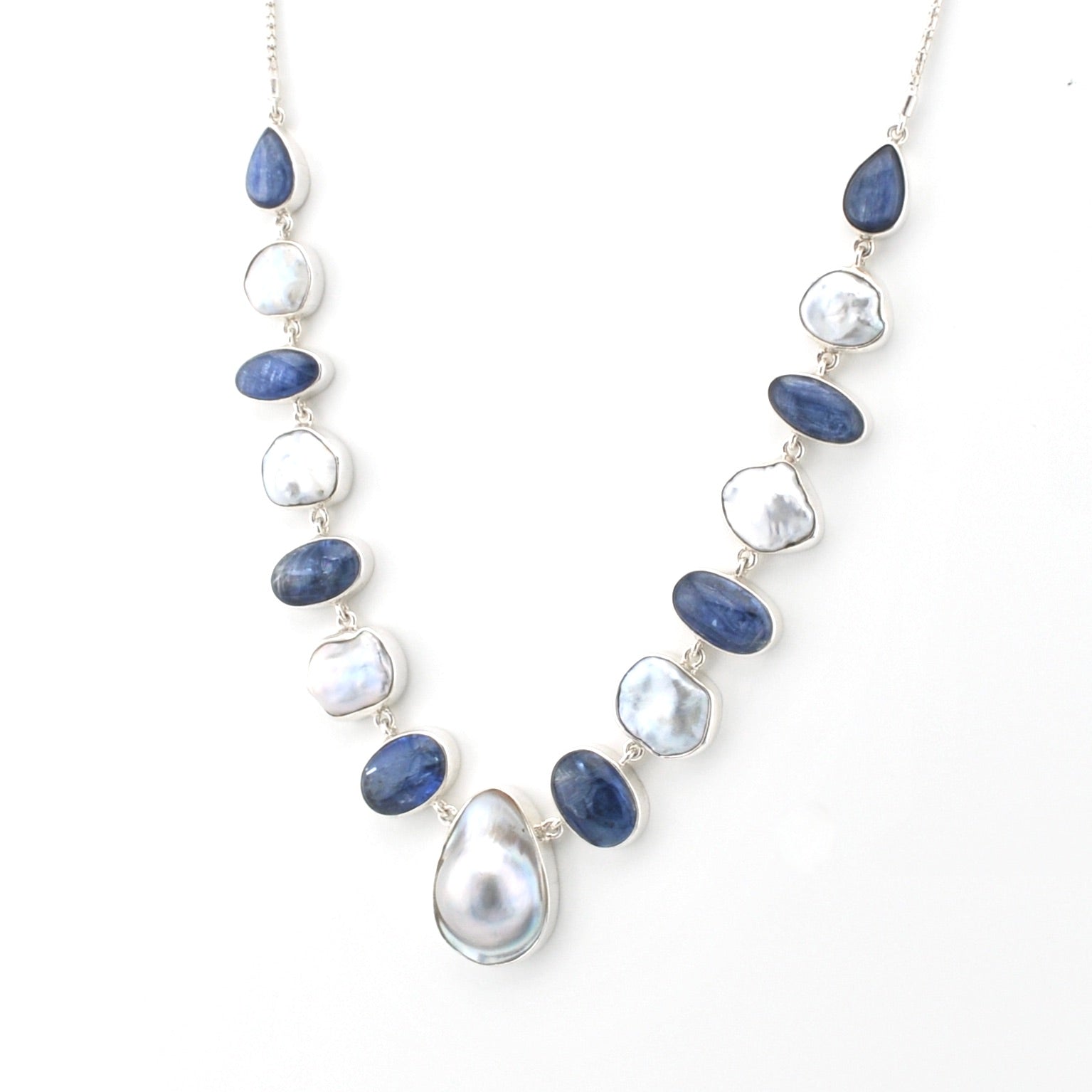 A silver-toned necklace featuring alternating polished dark blue sapphire-like beads and irregular white keshi pearl beads, with a large teardrop-shaped white pearl pendant at the center, displayed on a clean white background.