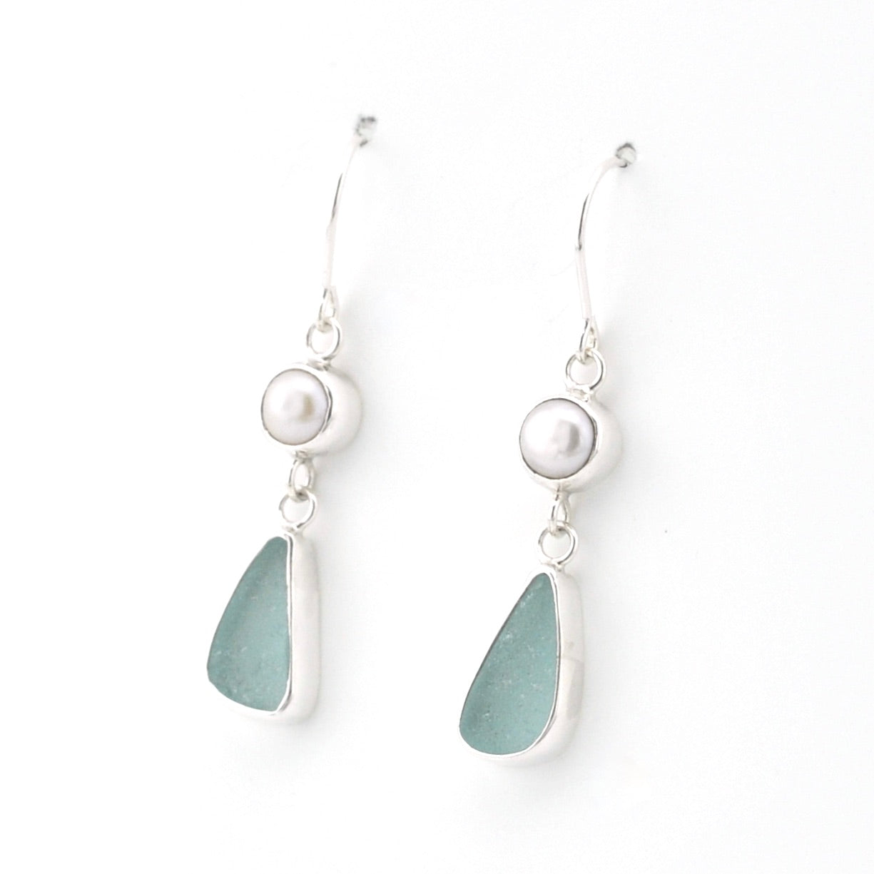 A pair of silver drop earrings featuring a small white pearl and a teardrop-shaped sea glass charm in a muted sage green color, set against a plain white background.