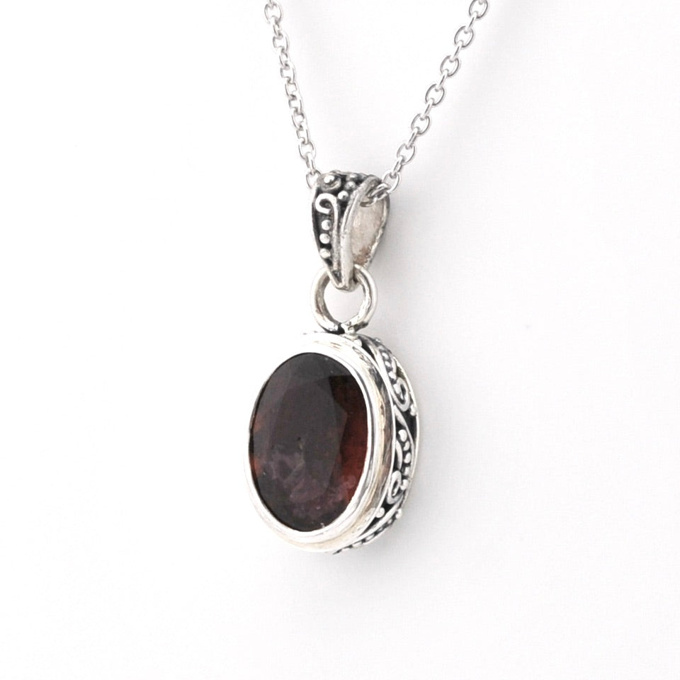 A silver chain necklace with an oval pendant featuring a dark reddish-brown gemstone in an ornate silver bezel setting, shown against a plain white background.