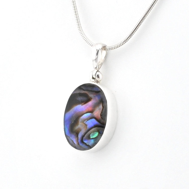 An oval pendant necklace featuring a silver bezel-set iridescent abalone shell inlay with blue, purple, and green hues, suspended from a silver snake chain against a plain white background.