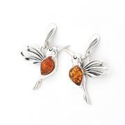 A pair of silver bird charms shaped like hummingbirds, each featuring a polished amber-colored heart-shaped stone in the center, set against a plain white background.