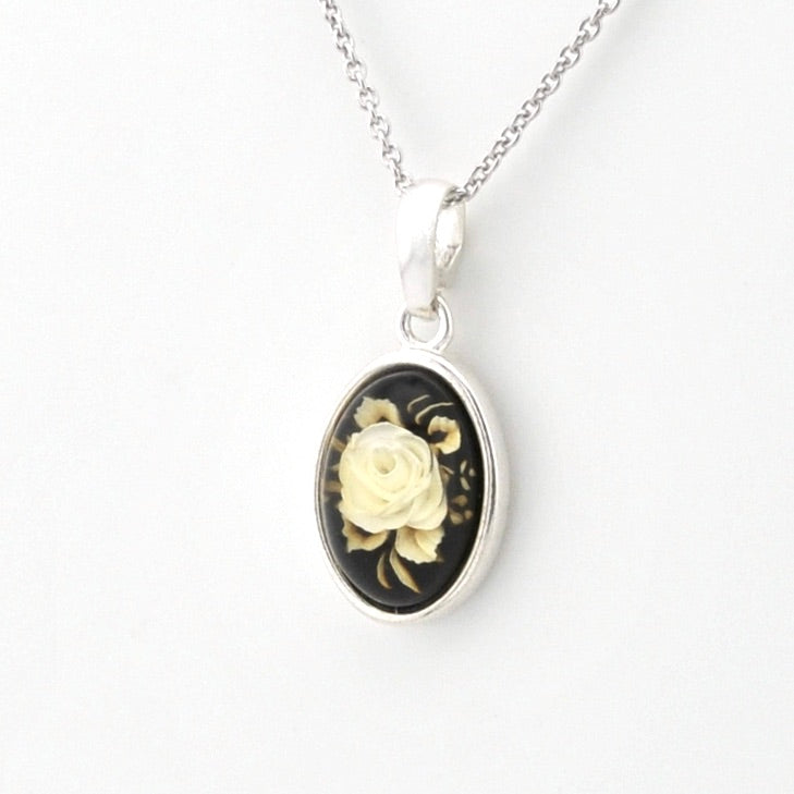 Silver oval pendant necklace featuring a black background with a detailed cream-colored rose and green leaves, suspended from a silver chain on a white background.