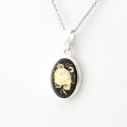 Silver oval pendant necklace featuring a black background with a detailed cream-colored rose and green leaves, suspended from a silver chain on a white background.
