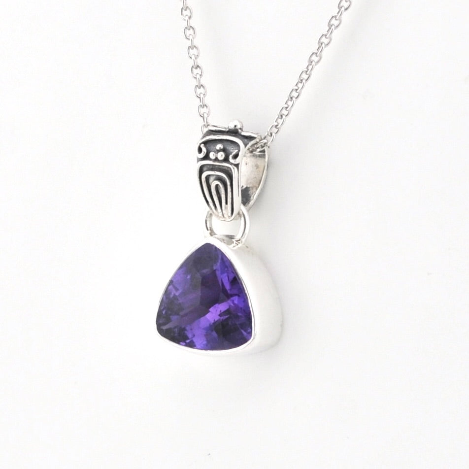 A silver pendant necklace featuring a triangular purple gemstone in a silver bezel setting, suspended from a silver chain with an ornate silver bail.