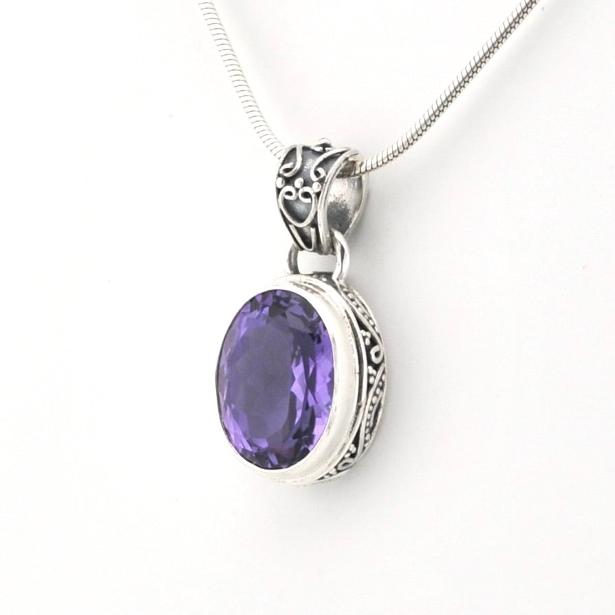A silver pendant necklace featuring an oval-cut purple gemstone in an ornate silver bezel setting, suspended from a silver snake chain against a plain white background.