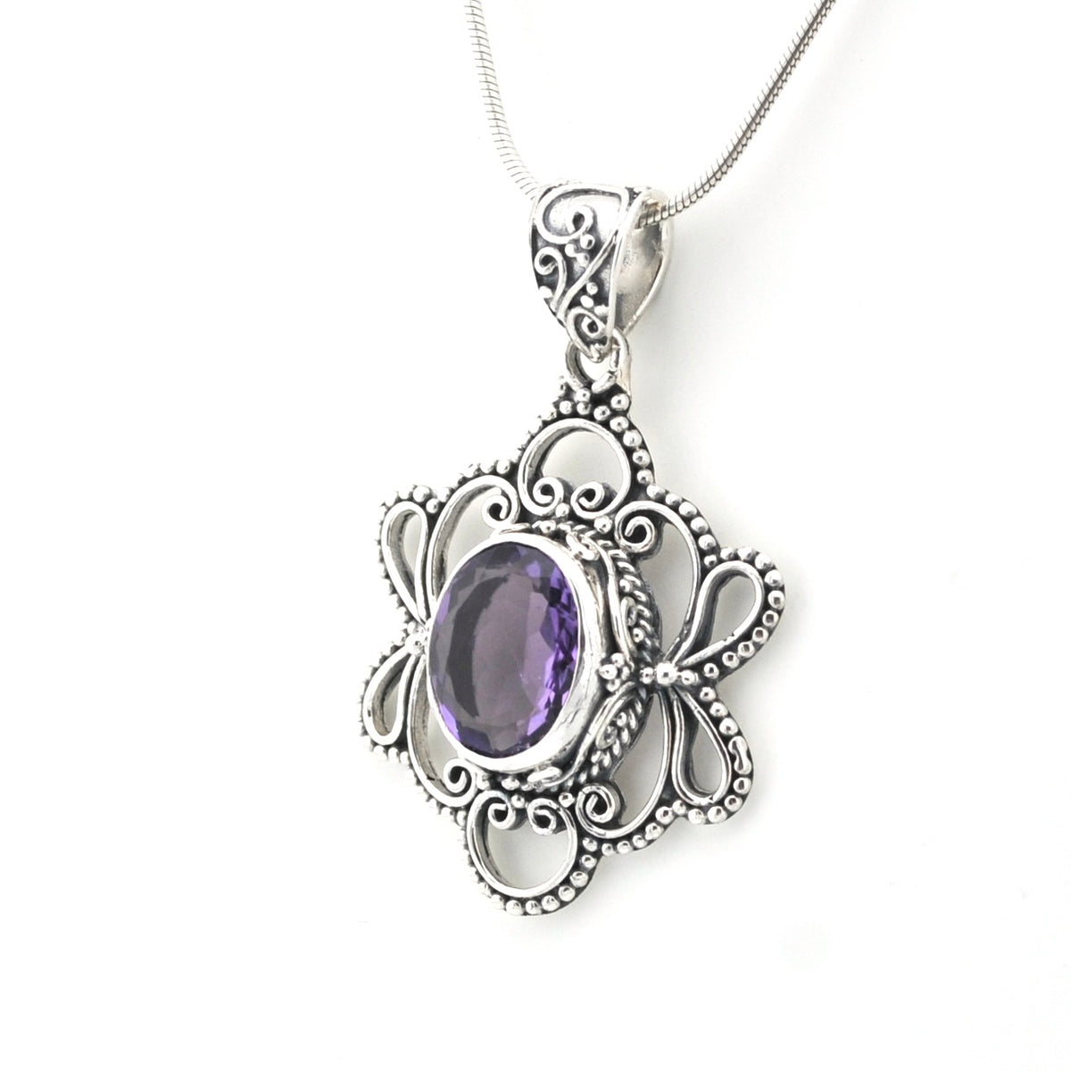 An ornate silver pendant necklace featuring a central oval purple gemstone surrounded by detailed filigree and beaded silver borders, suspended from a silver chain against a plain white background.