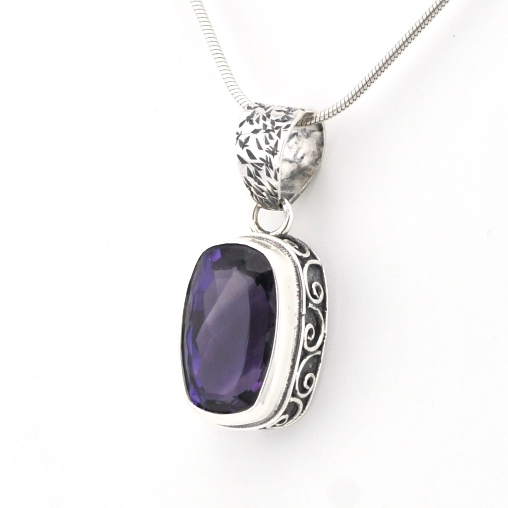 A silver pendant necklace featuring a rectangular, cushion-cut dark purple gemstone in an ornate, engraved silver bezel setting, suspended from a silver snake chain against a plain white background.