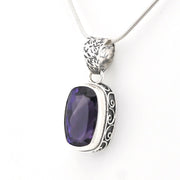 A silver pendant necklace featuring a rectangular, cushion-cut dark purple gemstone in an ornate, engraved silver bezel setting, suspended from a silver snake chain against a plain white background.