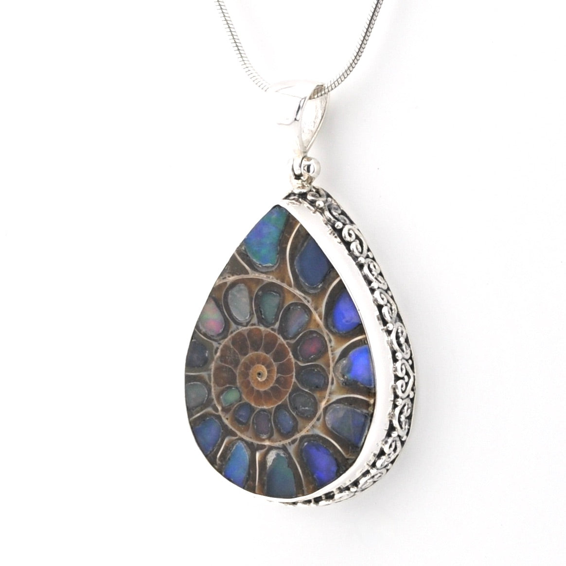 A teardrop-shaped pendant necklace featuring a fossilized ammonite shell pattern with iridescent blue, green, and brown hues, set in a silver-toned metal frame with an ornate side border and a silver chain.