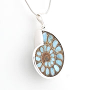 A silver pendant necklace featuring a polished ammolite shell inlay with light blue, brown, and tan concentric patterns, suspended from a silver snake chain against a plain white background.
