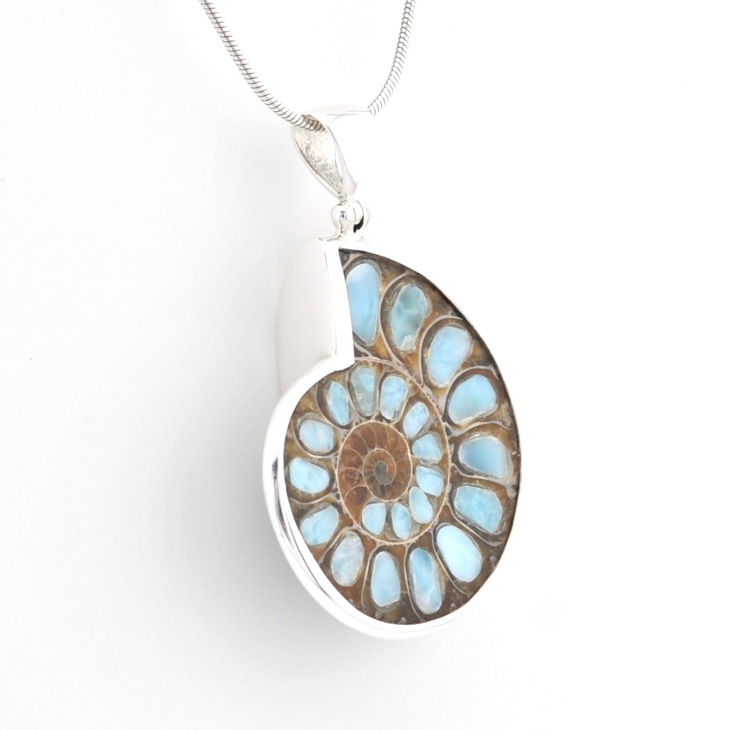 A silver pendant necklace featuring a polished ammolite shell inlay with light blue, brown, and tan concentric patterns, suspended from a silver snake chain against a plain white background.