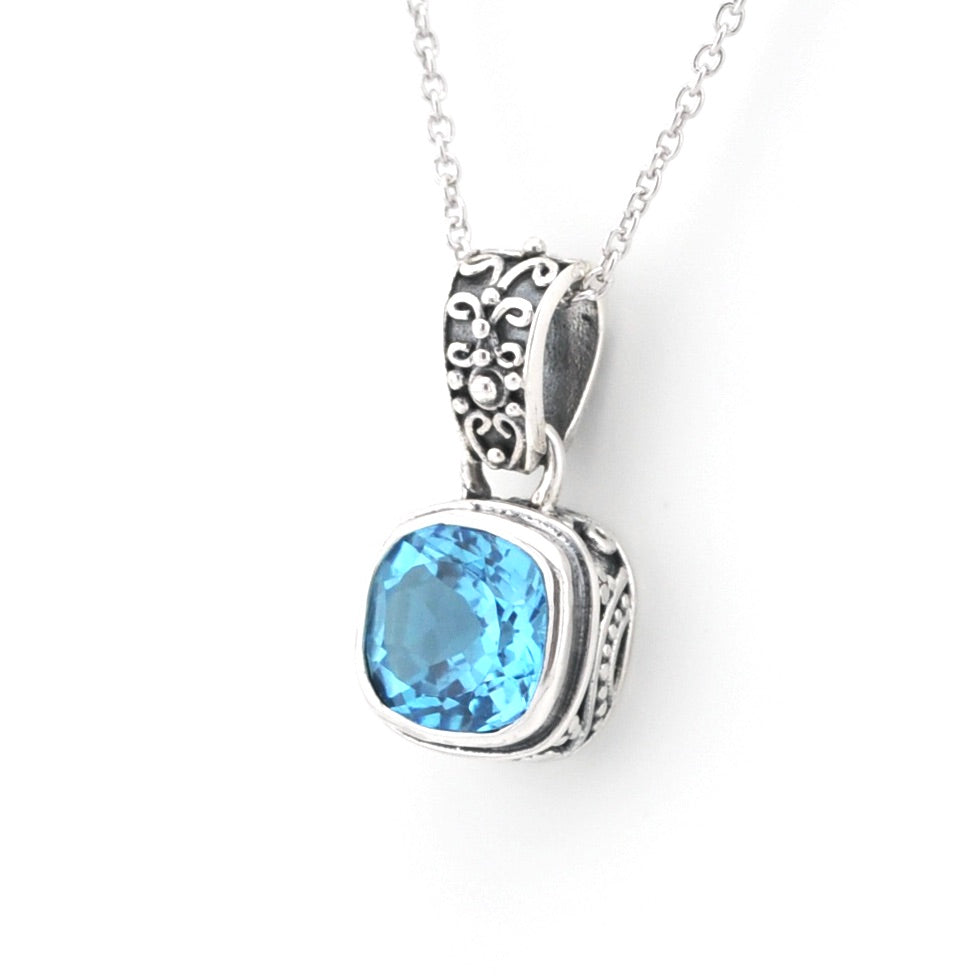 A silver pendant necklace featuring a cushion-cut light blue gemstone in an ornate, textured silver bezel setting, suspended from a delicate silver chain against a plain white background.