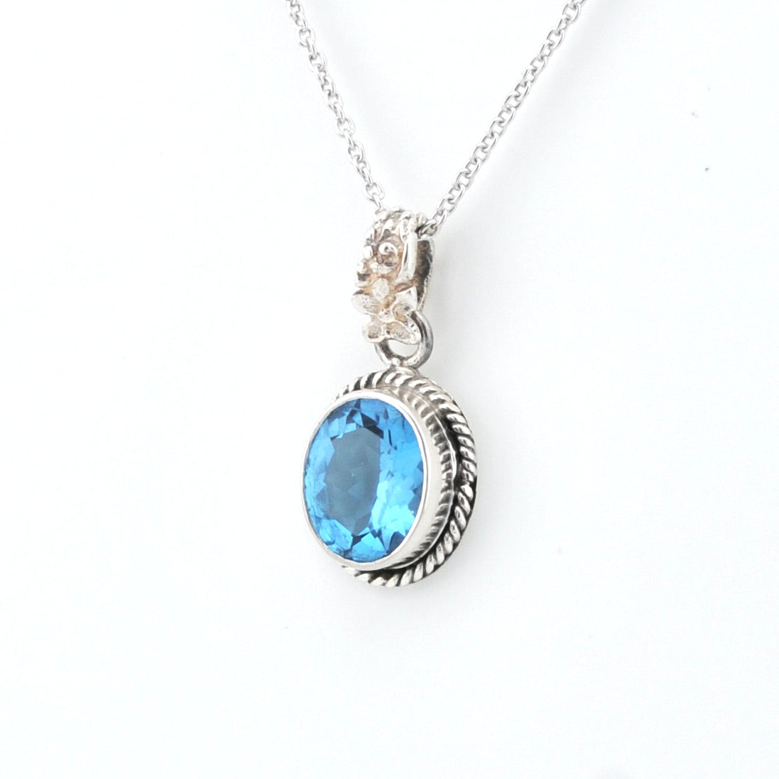A silver pendant necklace featuring an oval-cut light blue gemstone in a textured bezel setting, suspended from a delicate silver chain against a plain white background.